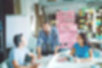 A group of people are gathered around a whiteboard, brainstorming ideas for app monetization. The whiteboard is filled with colorful sticky notes, flowcharts, and diagrams. Some people are holding cups of coffee, while others are pointing at different parts of the board. The atmosphere is lively and collaborative, with everyone contributing to the discussion. The focus is on finding creative ways to generate revenue from the app.